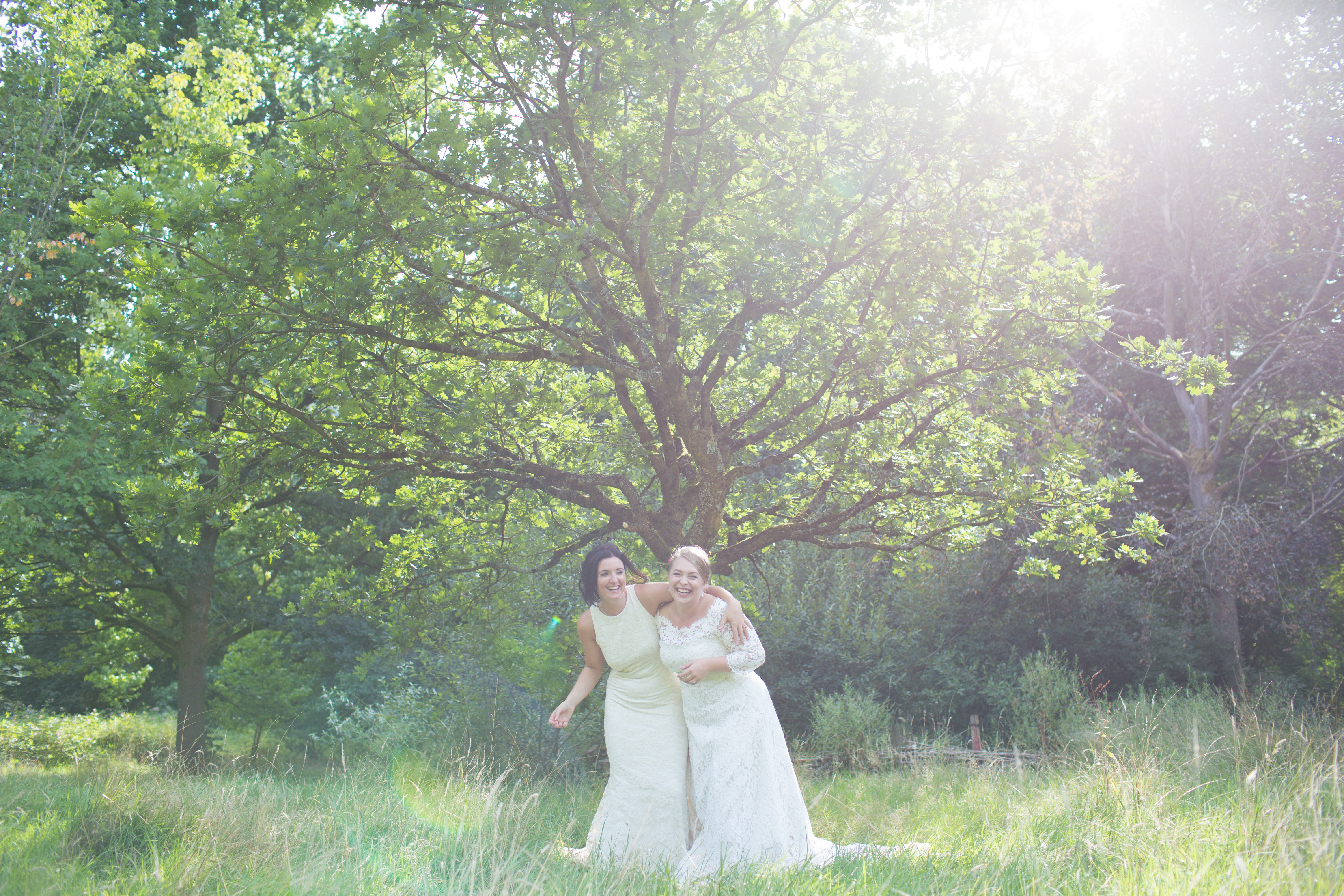 two brides laughing stood in front of a tree in the sun