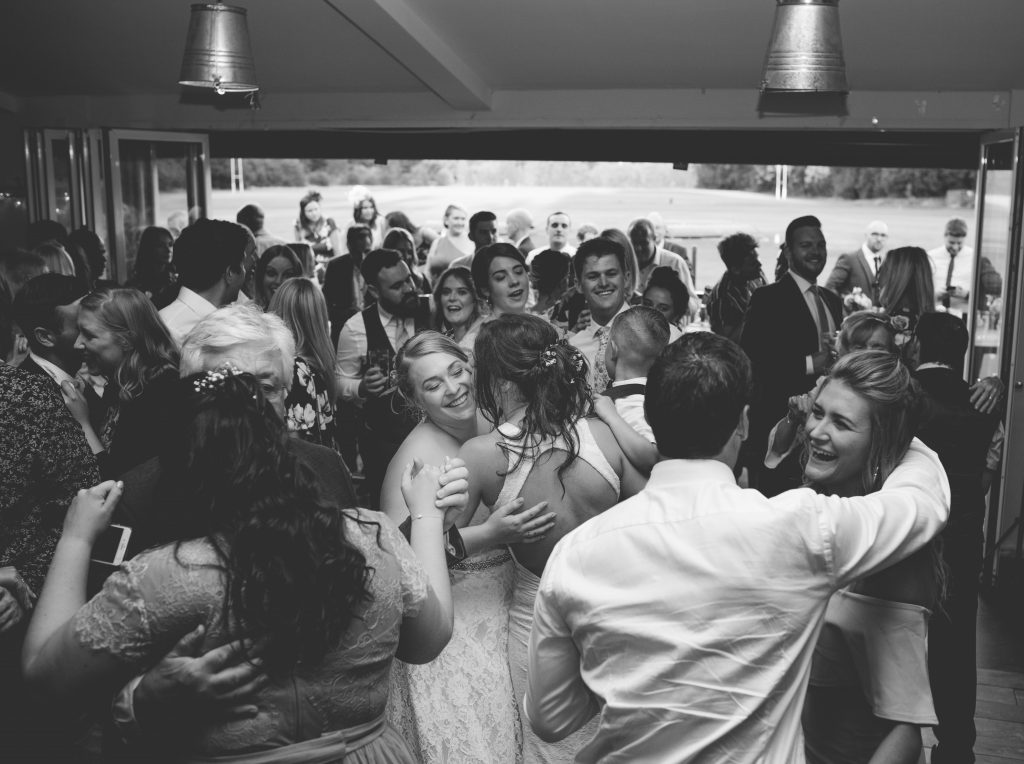 Black and white photo of two brides dancing surrounded by wedding guests