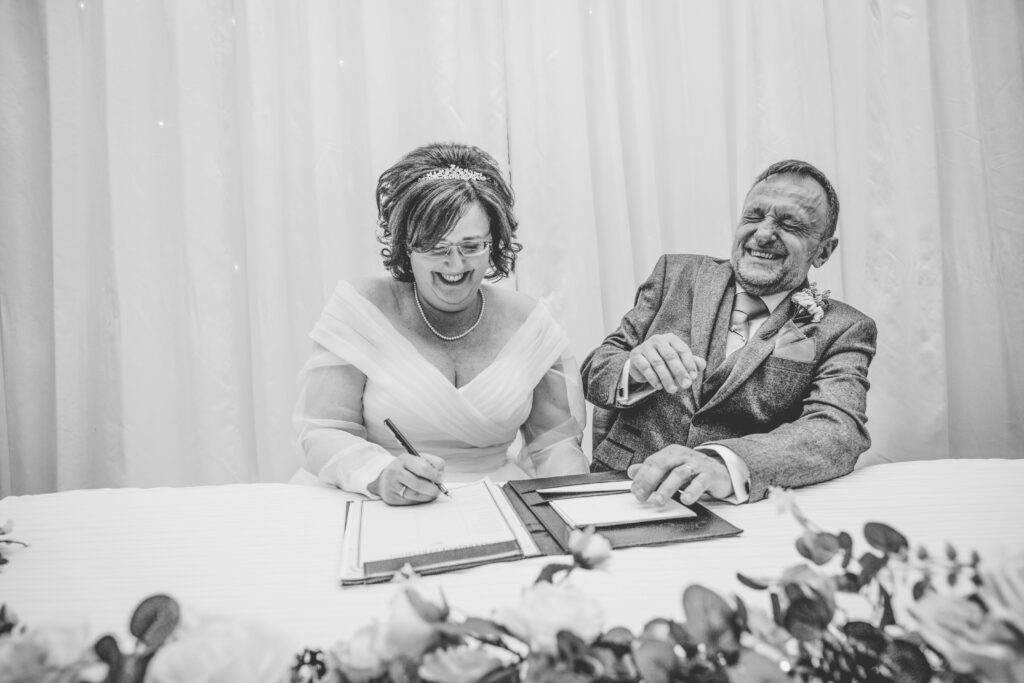 black and white photo of newly weds laughing whilst signing the wedding register